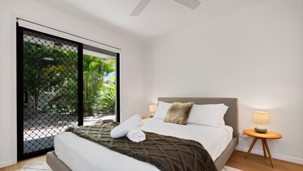 A bedroom with a bed, a nightstand, a lamp, and a view through a glass door. - Wyuna Drive 53 (Noosaville)