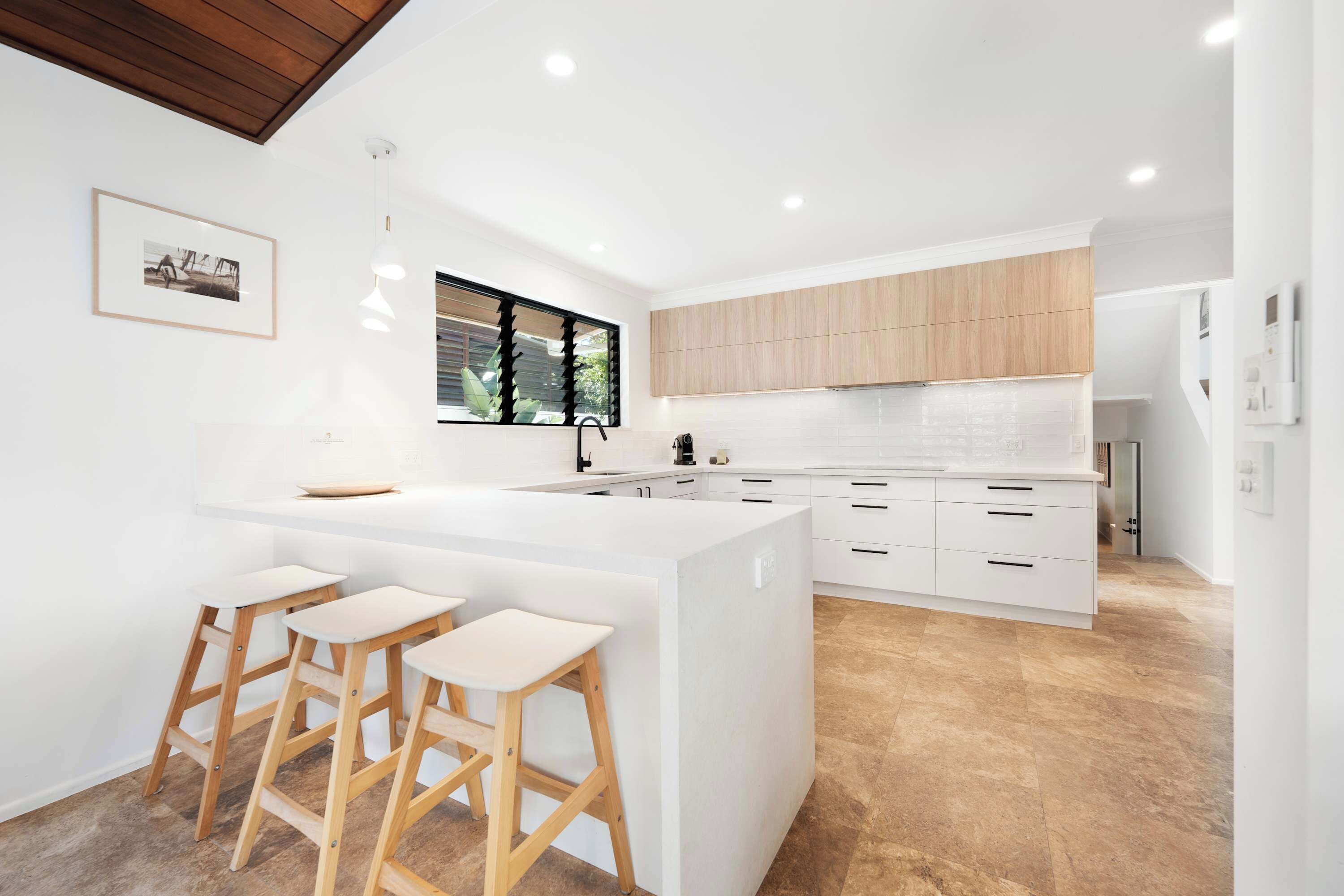 A modern kitchen with a central island, wooden stools, and a window with blinds.
