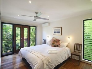 Une chambre dotée d’un grand lit, d’un ventilateur de plafond, d’une fenêtre avec des stores et offrant une vue sur la verdure environnante.