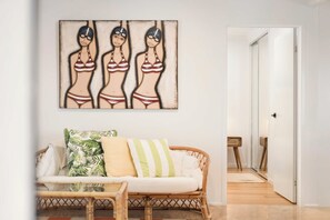 Une pièce avec un mur blanc, un canapé en osier agrémenté de coussins à rayures et un tableau représentant trois personnages.