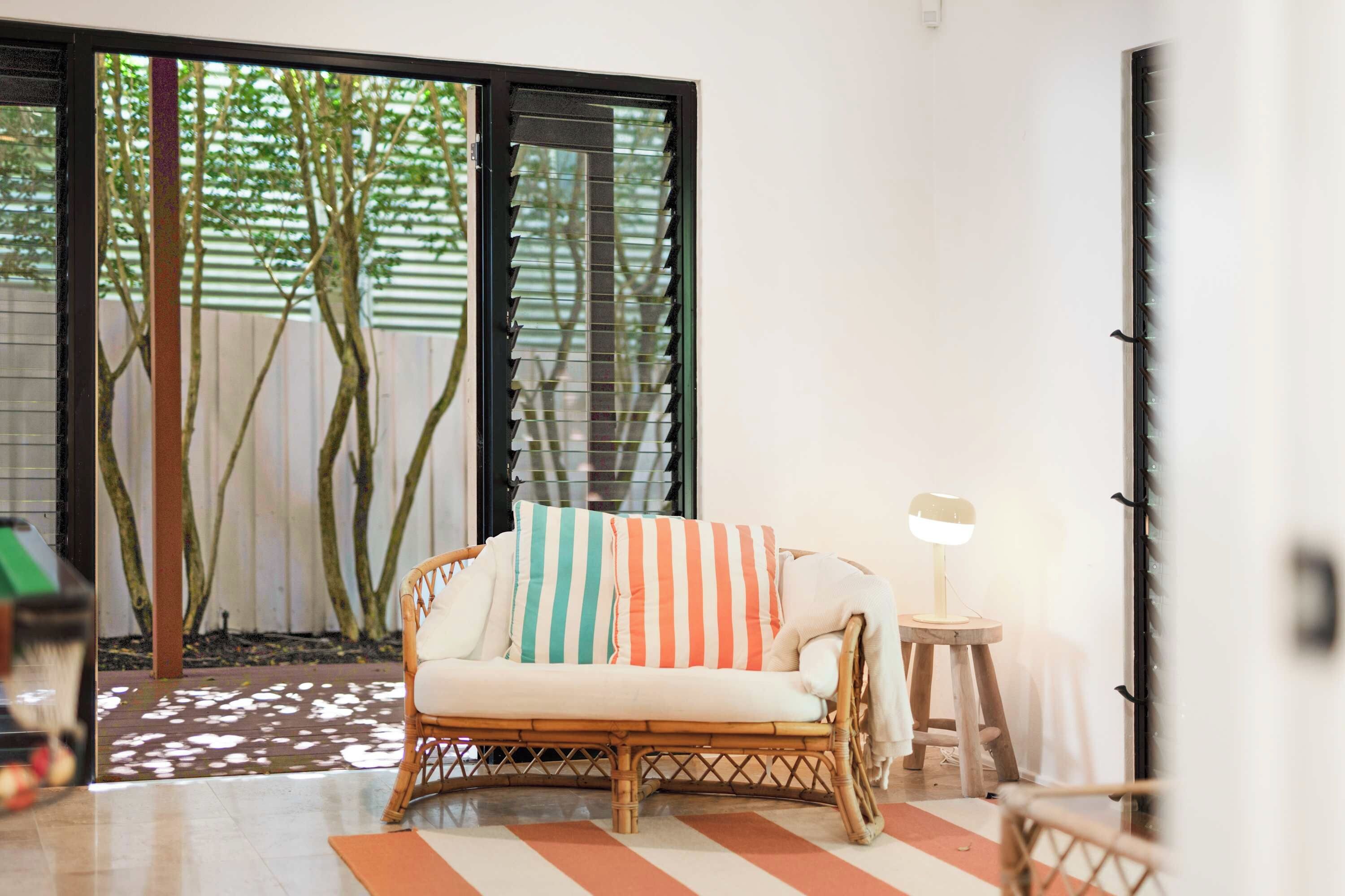 A wicker sofa with striped cushions, a small side table with a lamp, and a view of a garden through a glass door.