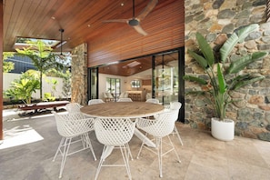 An outdoor dining area with a round wooden table and white lattice chairs, surrounded by stone walls and potted plants.