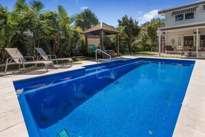 A rectangular swimming pool with clear blue water, surrounded by lounge chairs and a wooden gazebo.