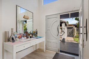Un meuble-lavabo blanc avec un grand miroir, une porte donnant sur l’extérieur et divers objets sur le comptoir, notamment des livres et des bouteilles.