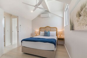 Un dormitorio con cama, ventilador de techo, aire acondicionado, lámparas de noche y un cuadro en la pared.