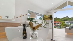 Et moderne interiør med et rundt bord, en flaske og glas, en dekorativ vase med planter, og udsigt til et boligområde gennem et vindue.