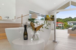 Un interior moderno con una mesa redonda que muestra una botella y copas, un jarrón decorativo con plantas, y vistas a una zona residencial a través de una ventana.