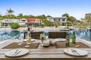 Mesa de comedor con platos, cubiertos y botellas, con vista a una piscina y a las casas y árboles circundantes.