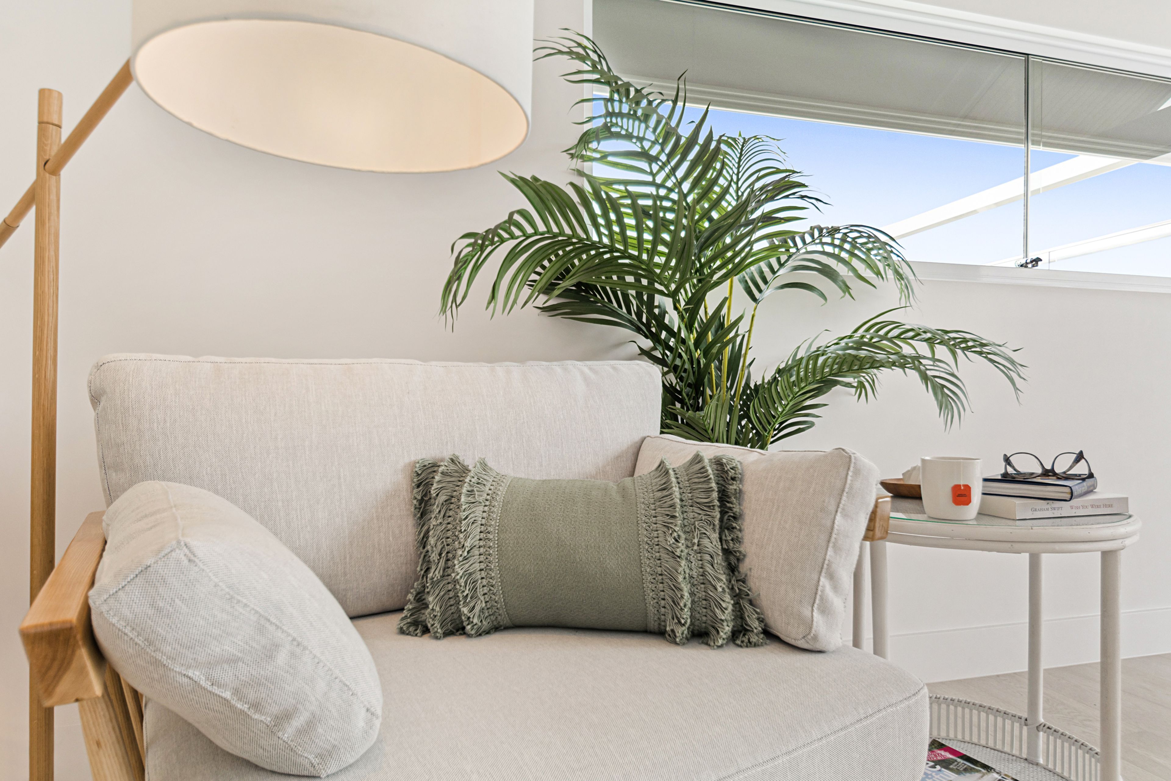 Ein modernes Wohnzimmer mit einer Couch, einer Stehlampe, einem kleinen Tisch mit Büchern und einer Tasse sowie einer großen Topfpflanze.