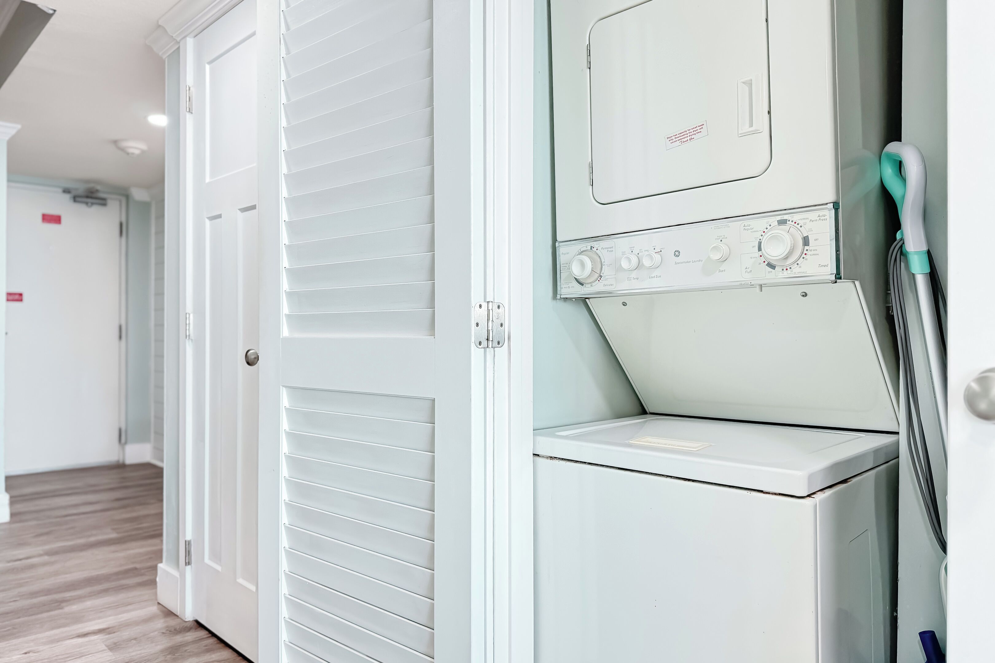 A laundry area with a washer and dryer, a door with louvered panels, and a hallway with wooden flooring.