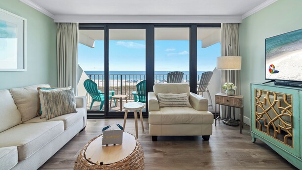Un salon comprenant un canapé, un fauteuil et une table basse, un téléviseur, ainsi qu’un balcon avec des chaises et une vue sur la plage.