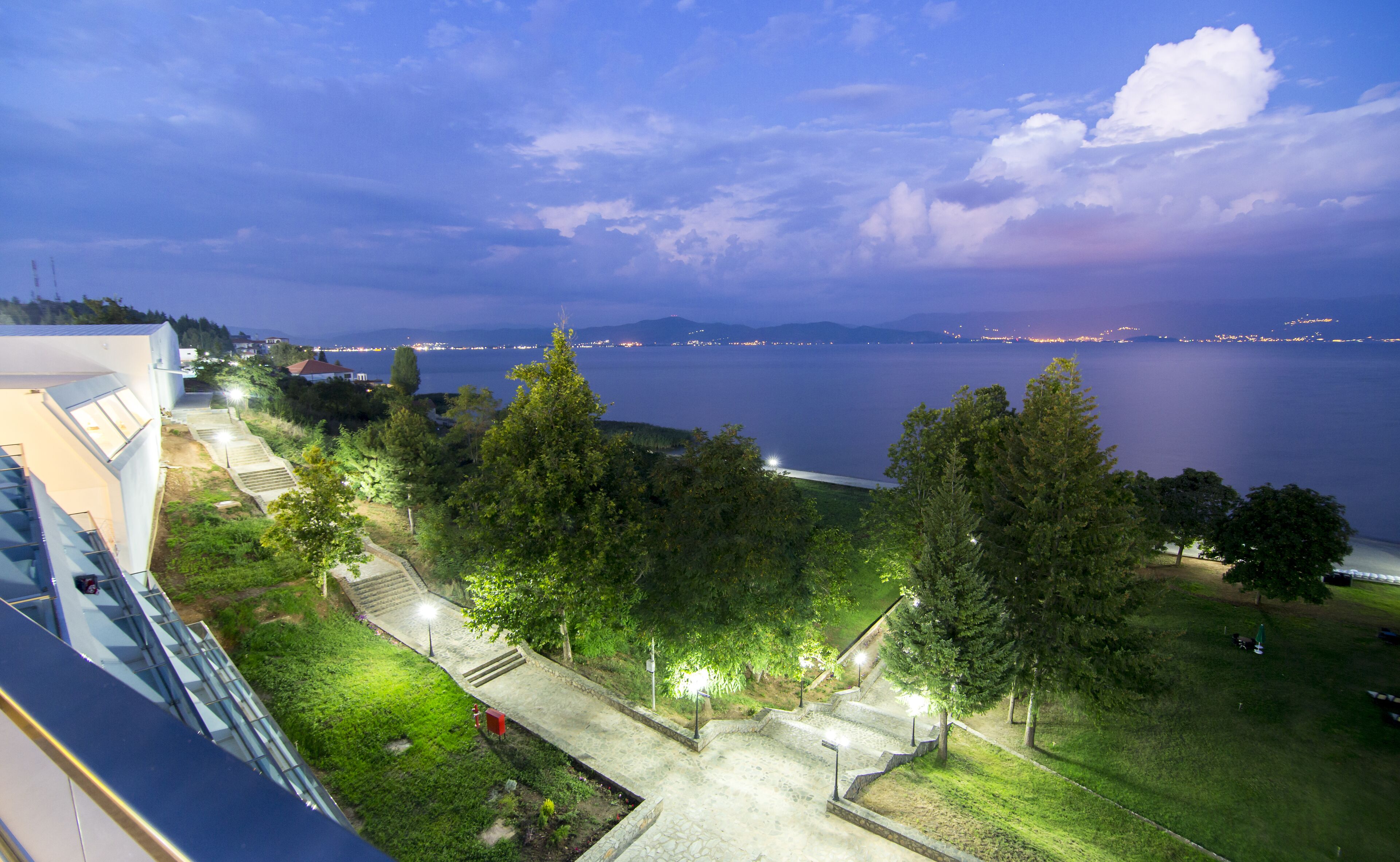 A coastal view with a pathway leading to a grassy area, overlooking a body of water and distant city lights.