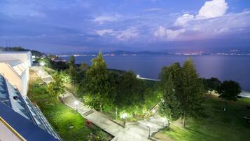 Vista costera con un camino que lleva a una zona verde, con vista a una masa de agua y luces de la ciudad a lo lejos.