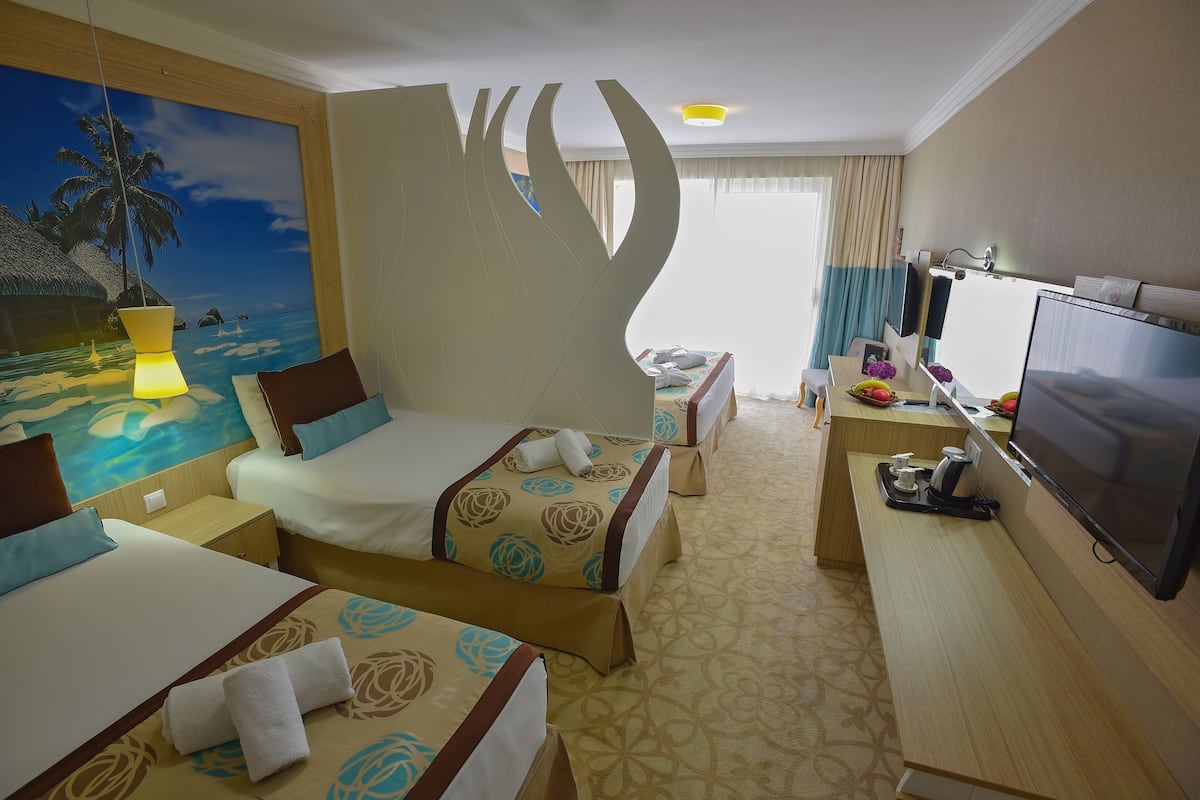 A hotel room with two beds, a desk, a TV, and a large window with curtains.