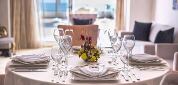 Una mesa de comedor preparada para seis personas, con cristalería y platos transparentes, un centro de mesa con flores y vista al mar a través de grandes ventanales.