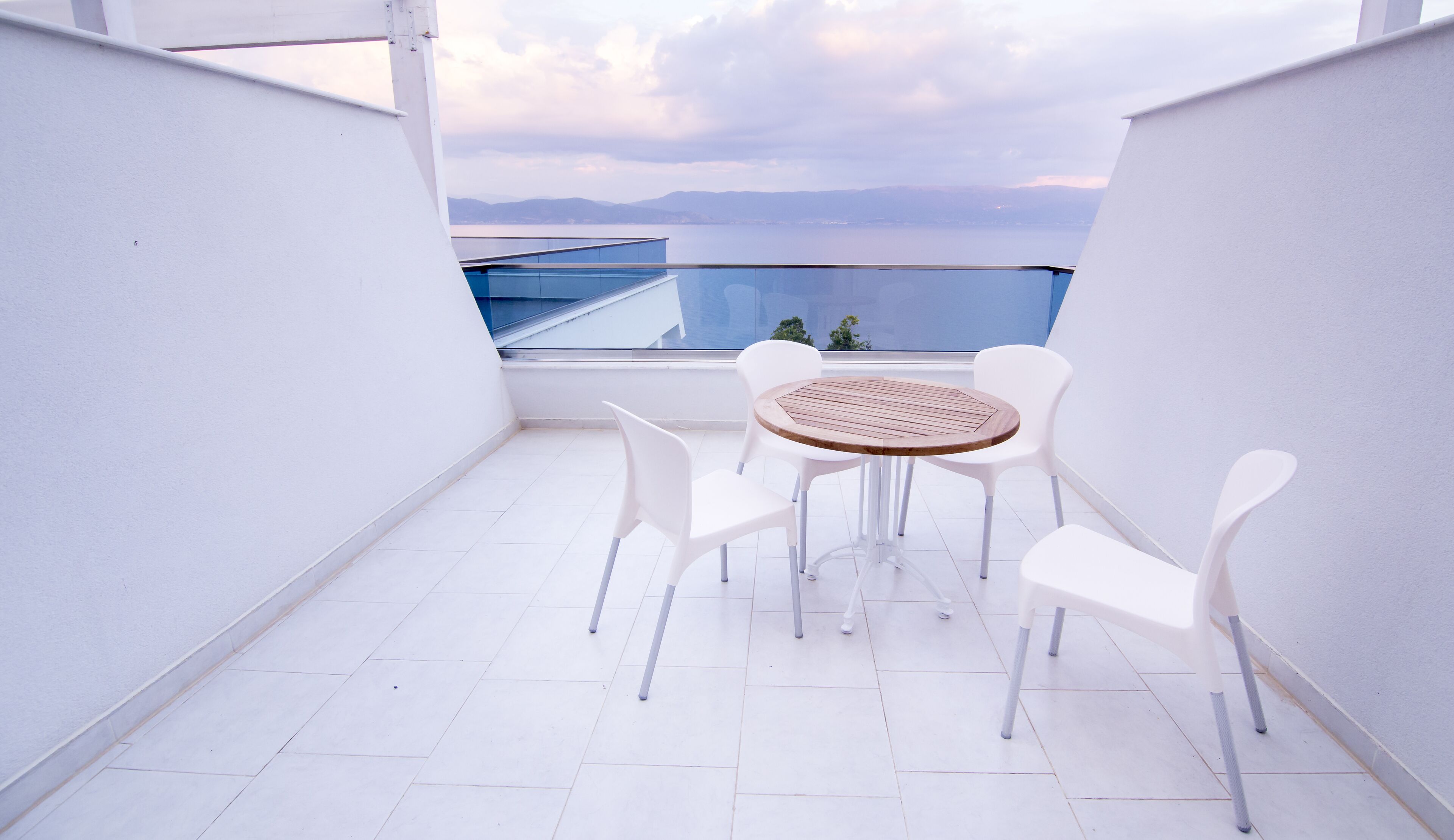 Un balcone con un tavolo rotondo in legno e sedie bianche, con vista su un corpo d'acqua e montagne lontane.