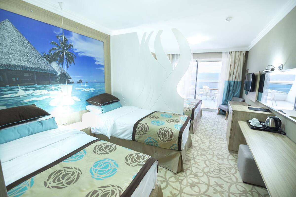 A hotel room with two beds, a desk, a TV, and a large window with a view of the ocean.