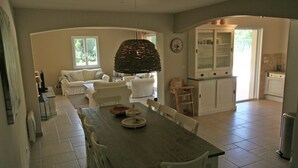 A spacious living area with a dining table, white chairs, a sofa, and a kitchen area with cabinets and a sink.