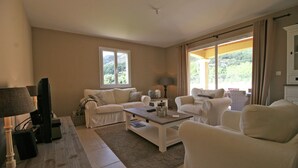 A living room with a sofa, armchairs, a coffee table, and a TV. There is a window with curtains and a view of greenery outside.