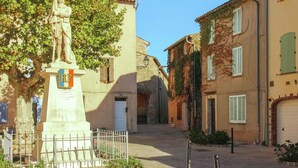 A narrow street with a statue, buildings with shutters, and a clear sky.