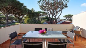 Ein Tisch mit Glasplatte, Stühle, ein Korbwäsche und eine Schale mit Früchten und Blumen auf einer Terrass mit Fliesenboden und Blick auf Bäume und Gebäude.