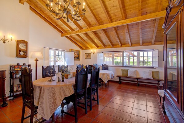 A dining area with a wooden ceiling, a chandelier, a table with chairs, a bench, and windows with a view.