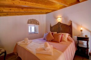 A bedroom with a wooden ceiling, a large bed with a headboard, two wall-mounted lights, and a small table with a lamp.