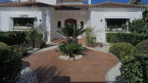 Un edificio blanco con tejado de tejas, una entrada central con arco y un patio empedrado con plantas.