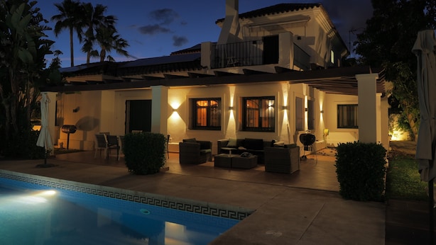 Una casa de dos plantas con piscina, mobiliario de exterior y un patio bien iluminado.