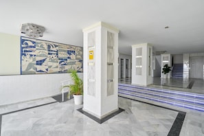A lobby with a tiled wall mural, a potted plant, a white pillar, and a staircase with blue accents.