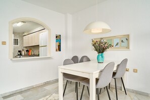 Un comedor con una mesa blanca, sillas grises, un florero con flores y una lámpara fijada a la pared.
