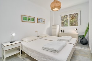 Un dormitorio con cama, mesitas de noche, lámpara, ventana con vistas y cuadros en la pared.
