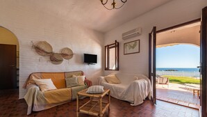Ein Zimmer mit weißer Ziegelwand, Fernseher, Sofa, kleinem Tisch und Meerblick durch eine geöffnete Tür.