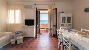 A room with a TV, a dining table with a bowl and plates, wicker baskets, a bed, and a view of the beach. - Holiday Home "Nicoleup" with Sea View, Terrace & Shared Garden (Perd'e Sali)