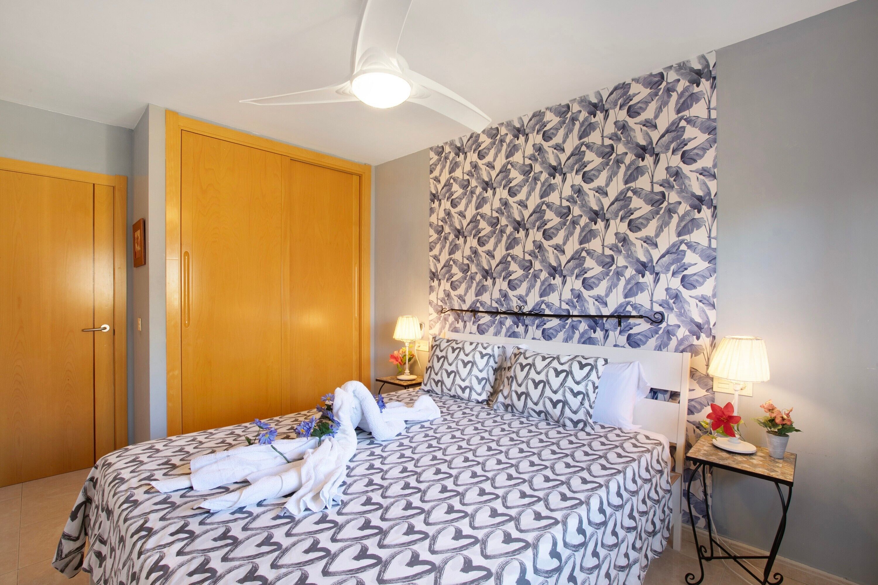 A hotel room with a bed, bedside tables, a ceiling fan, and a wall with a leaf pattern.