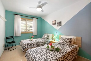 A room with two beds, a ceiling fan, a window with curtains, and a small table with a lamp.