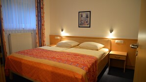 Habitación de hotel con cama, mesita de noche, cajón, un cuadro enmarcado en la pared y ventana con cortinas.