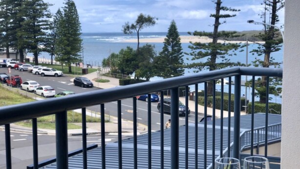 A balcony with a view of a beach and ocean, a parking lot with cars, and a pedestrian pathway. - Grand Pacific U55 | Private Spa Balcony, Water Views & Walk To Beach (Caloundra)