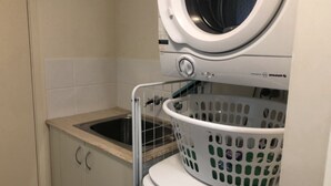 A laundry area with a washing machine, a sink, and a laundry basket. - Grand Pacific U55 | Private Spa Balcony, Water Views & Walk To Beach (Caloundra)