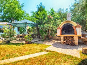 Uma área de pátio externa com um forno a pizza de tijolos, um espaço coberto com cadeiras e um caminho de pedra.