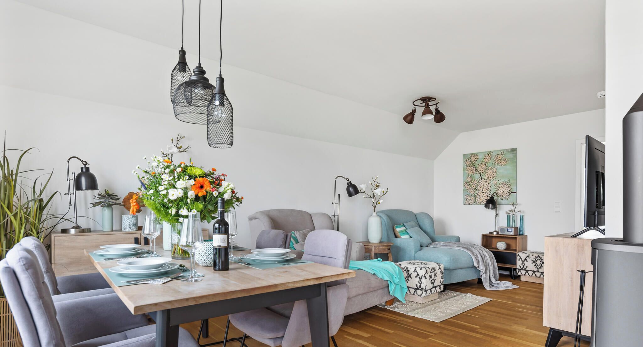 A modern living room with a dining area, featuring a wooden dining table, grey sofas, and a grey couch. There are hanging lights, a TV, and a painting on the wall.