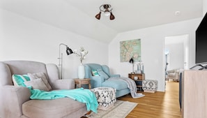 A modern living room with a grey sofa, a blue armchair, and a patterned ottoman. There is a floor lamp, a vase with flowers, and a painting on the wall.
