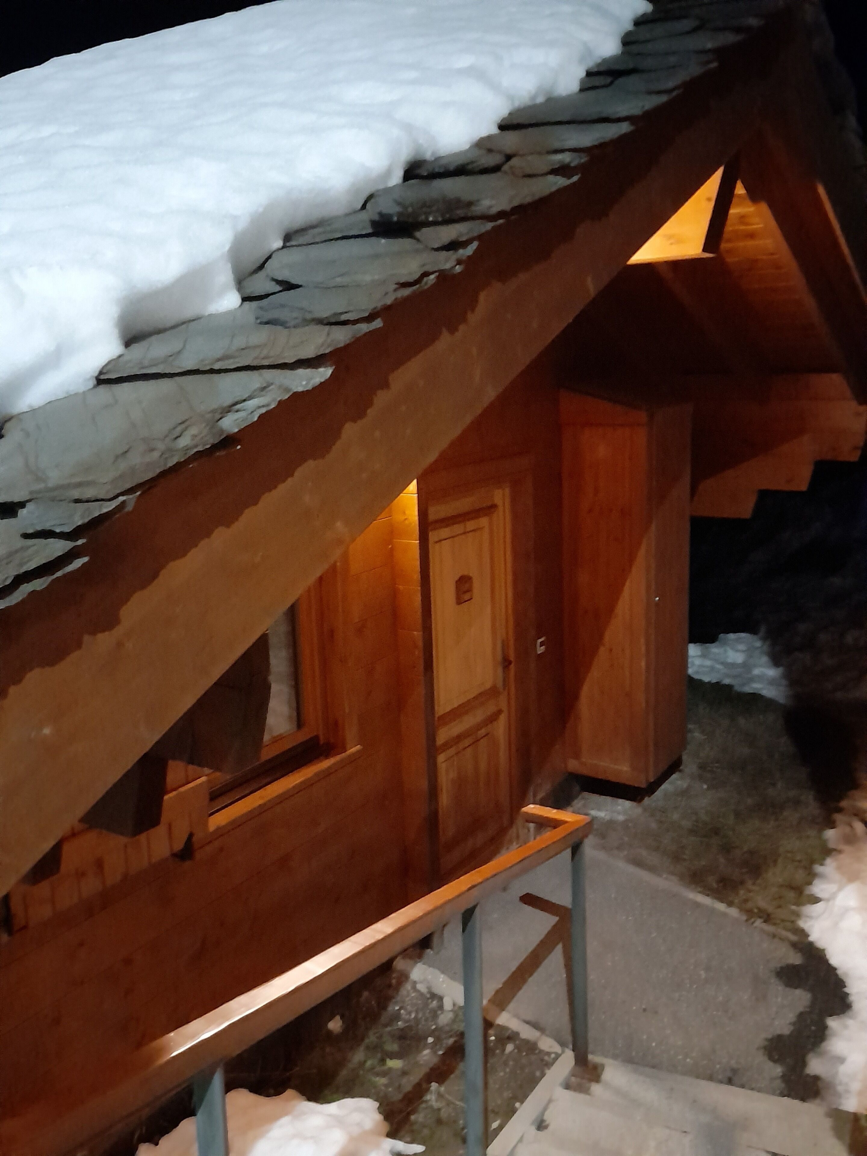 Een houten chalet met een sneeuwbedekte, schuine dak en een houten deur, met een klein terras.