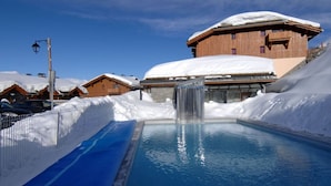 A pool with a waterfall, surrounded by snow-covered buildings and a clear sky.