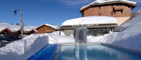 A pool with a waterfall, surrounded by snow-covered buildings and a clear sky.
