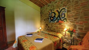 A room with a stone wall, a bed with floral bedding, a nightstand with a lamp and flowers, and a wooden cabinet. - Belvilla by OYO Villa Lucignano (Lucignano)