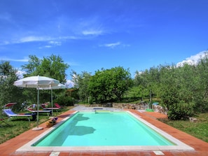 A rectangular swimming pool with a tiled border, surrounded by a brick patio. There are lounge chairs and a white umbrella in the background.