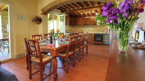 A dining area with a wooden table and chairs, a kitchen in the background, and a vase of purple flowers on the table. - Belvilla by OYO Villa Lucignano (Lucignano)