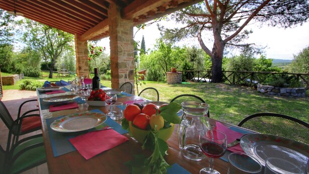 An outdoor dining area with a table set for two, featuring a bowl of fruit, wine glasses, and a pitcher. - Belvilla by OYO Villa Lucignano (Lucignano)