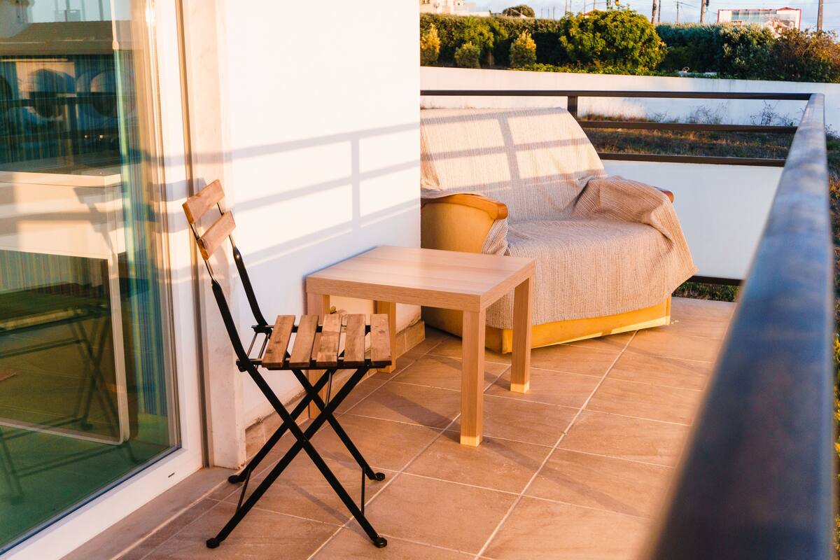 Een balkon met een houten stoel, een tafeltje en een bank met een plaid.